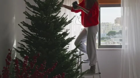 Woman on ladder wrapping string lights around Christmas tree at home, 4K Stock Footage 323967862