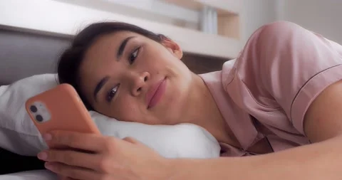 Woman laughs after reading a message on her mobile phone while lying in bed Stock Footage 184283380