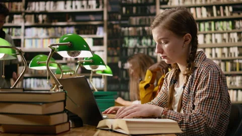 Woman learner studying at shared library table with laptop and open book Stock Footage 337229352