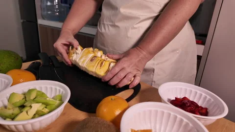 A woman in a light apron is sorting a jackfruit into slices, there are many Stock Footage 329936058