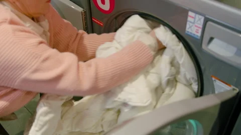 Woman loading a large white comforter into a front-loading washing machine at a Stock Footage 328105008