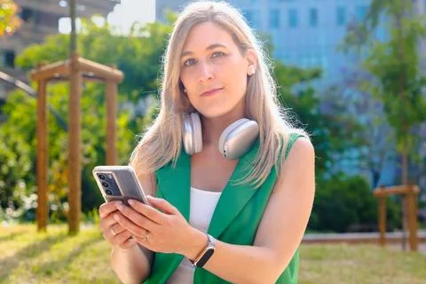 Woman looking at camera while holding smartphone outdoors in sunny park Stock Photos