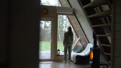 A woman looking through a panoramic window of a wooden house with a fireplace. Video stock 322507770