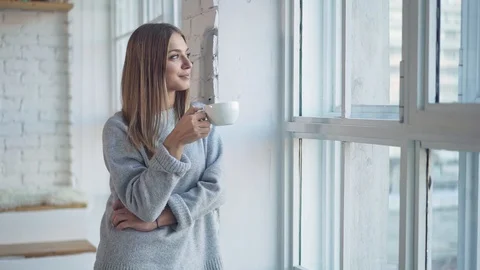 Woman looking in window and drinking tea from white cup Stock Footage 100041843