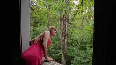 Woman looks through an open window frame toward a forest Stock Footage 346076561