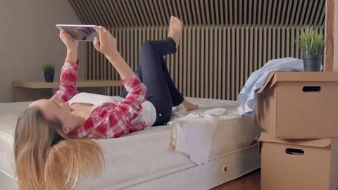 Woman lying on bed using digital tablet instead of unpacking boxes of clothes in Stock-Footage 72231893
