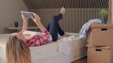 Woman lying on bed using digital tablet instead of unpacking boxes of clothes in Stock Footage 72232048