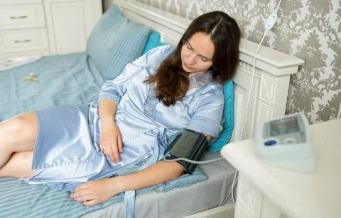 Woman lying on bed using electronic blood pressure monitor, relaxing during h Stock Photos