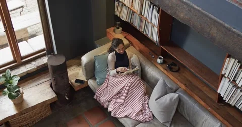 Woman lying on sofa under blanket and reading book at home, slow motion Stock Footage 344849748