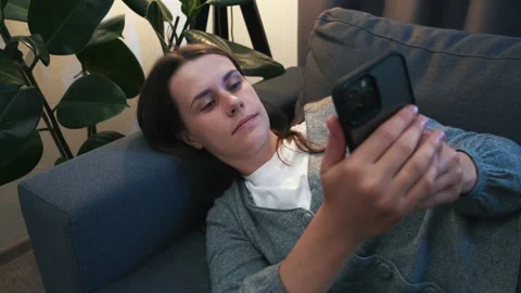 Woman lying on sofa under light lamp browsing surfing wireless internet on phone Stock Footage 202123002