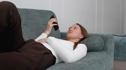 Woman lying on sofa using smartphone, changing facial expressions Stockbeeldmateriaal 308318150