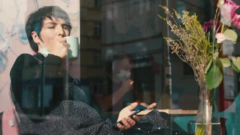 Woman in mask drinking tea while sitting by window in cafe. Distance COVID-19. Stock-Footage 142148932