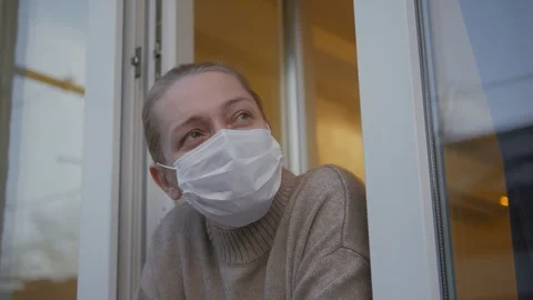 A woman in a medical mask looks out of the window and removes the mask. Stock Footage