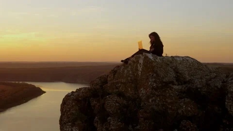 A woman opens a laptop while sitting on a rock above the river at sunset Stock Footage 234697499