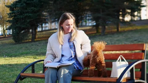 A woman is peacefully reading a book while sitting on a bench with her dog in Stock Footage 307817654