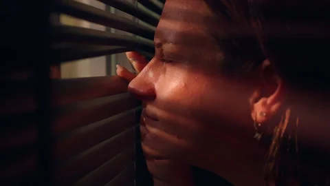 Woman Peeks Through Window Blinds in a Dark Interior, Shadow Stripes Convey Priv Stock Footage 345137116