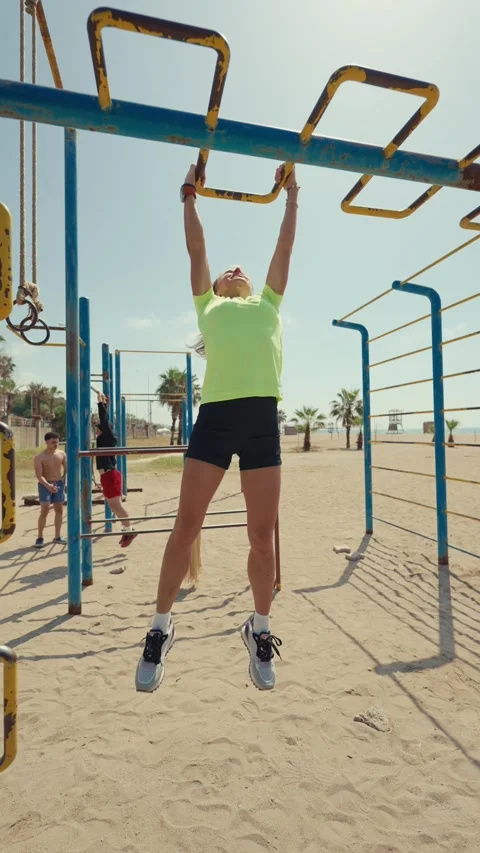 A woman performs pull-ups on monkey bars at an outdoor beach gym. The vertical Stock Footage 304687139