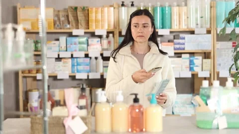 Woman in pharmacy scans QR code on emollient cream product, studies product in Stock Footage 325765875