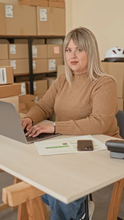 Woman pointing finger at camera while typing on laptop amid parcel boxes in.. Stock Footage 347850698