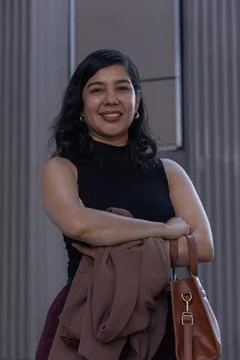 Woman posing with folded brown jacket, handbag; wearing black top, gold hoops at Foto stock