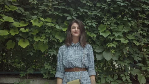 Woman posing in front of camera, standing by overgrown fence touching leafs Video stock 145991644