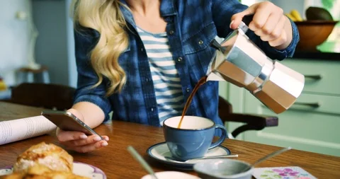 Woman Pouring Coffee While Checking Smart Phone At Breakfast Table Stock Footage 100039379