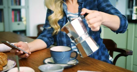 Woman Pouring Coffee While Checking Smart Phone At Breakfast Table Stock Footage 100039409