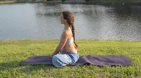 Woman practicing baddha konasana outdoors on the grass near lake Stock Footage
