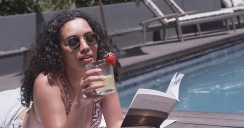 Woman is propping on arms while reading paperback by pool, reaching for drink to Stock Footage 337026119