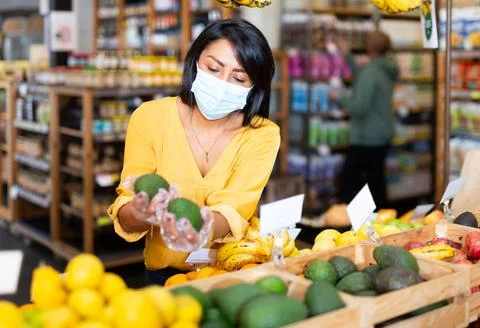 Woman in protective mask shopping in organic food store, choosing avocado Stock Photos