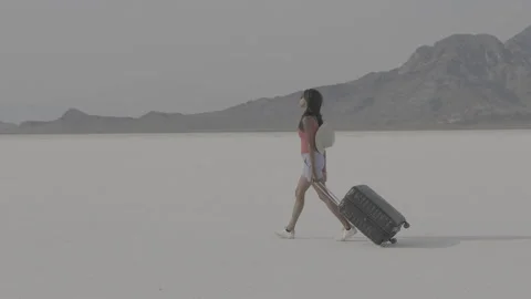 Woman pulling suitcase through vast desert landscape under bright sky Stock Footage 312646628