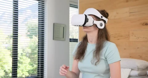 Woman reaching for white VR headset by phone on bed, adjusting and selecting Stock Footage 341665962