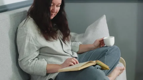 Woman reading book while drinking coffee on sofa at home Stock Footage 291497941