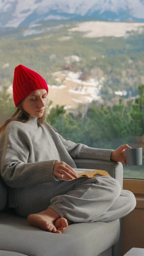 A woman reading by a large window and enjoying scenic snowy mountain view. 動画素材 343446773