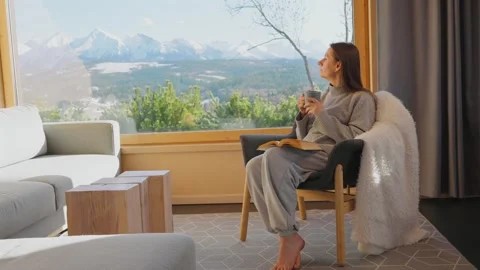 A woman reading by a large window and enjoying scenic snowy mountain view. Stockbeeldmateriaal 343446863