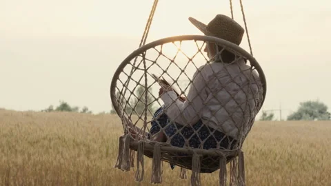 Woman reading a paper book while relaxing on a hanging swing beside a golden Stock Footage 340023827