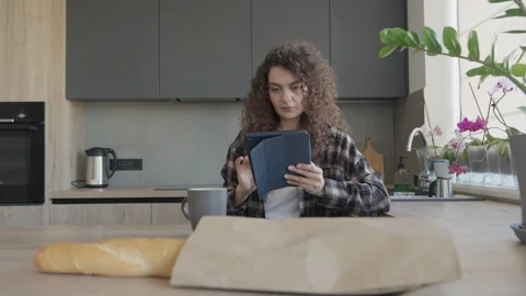 Woman Reading On Tablet While Sitting In Kitchen With Groceries And Mug Nearby Stock Footage 306396377