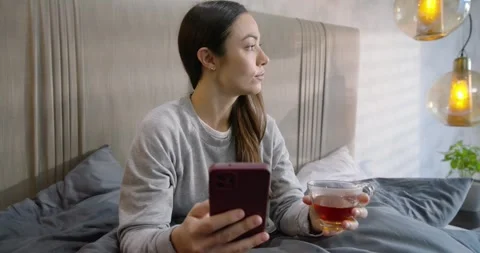 Woman receiving notification, checking smartphone, smiling and sipping tea from Stock Footage 339231875