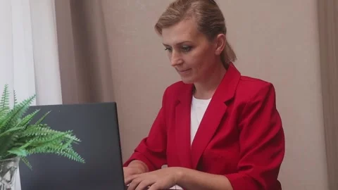 Woman in a red jacket sits at the table typing text on a laptop smiling friendly Stock Footage 251641199