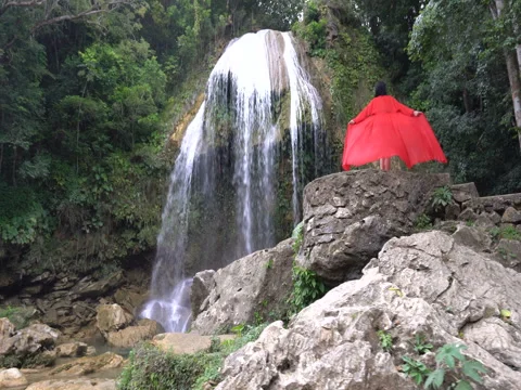 Woman in red long fluttering dress is standing near waterfall in tropics Stock Footage