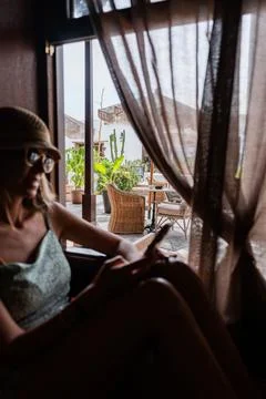Woman relaxes indoors while using smartphone near window with greenery view Stock Photos