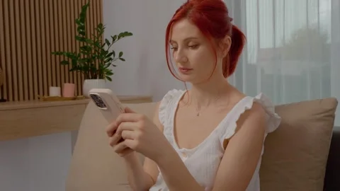 Woman relaxes on sofa while browsing her phone in her living room Stock Footage 329231687