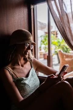 Woman relaxing by the window while using her smartphone during sunny day Stock Photos