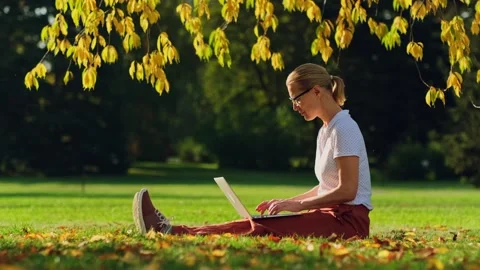 Woman remotely working while sitting on the lawn in autumn park. Видео 251518601