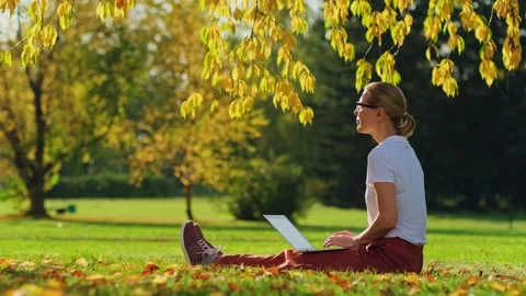 Woman remotely working while sitting on the lawn in autumn park. Stock Footage 274250304
