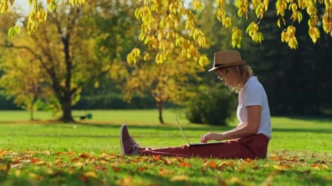 Woman remotely working while sitting on the lawn in autumn park. Stock Footage 278379442