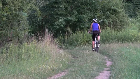 A woman rides a bicycle on a path along the edge of a forest Stock Footage