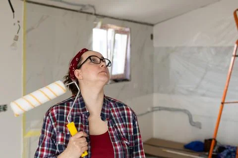 A woman with a roller in hands looks up while examining a surface for painting. Stock Photos