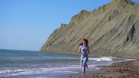 A woman runs along a sandy beach with turquoise water against a rocky shore Stock Footage 142399341