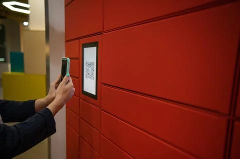 A woman scans a red code to pick up a parcel at a parcel machine. Automated Stock Photos